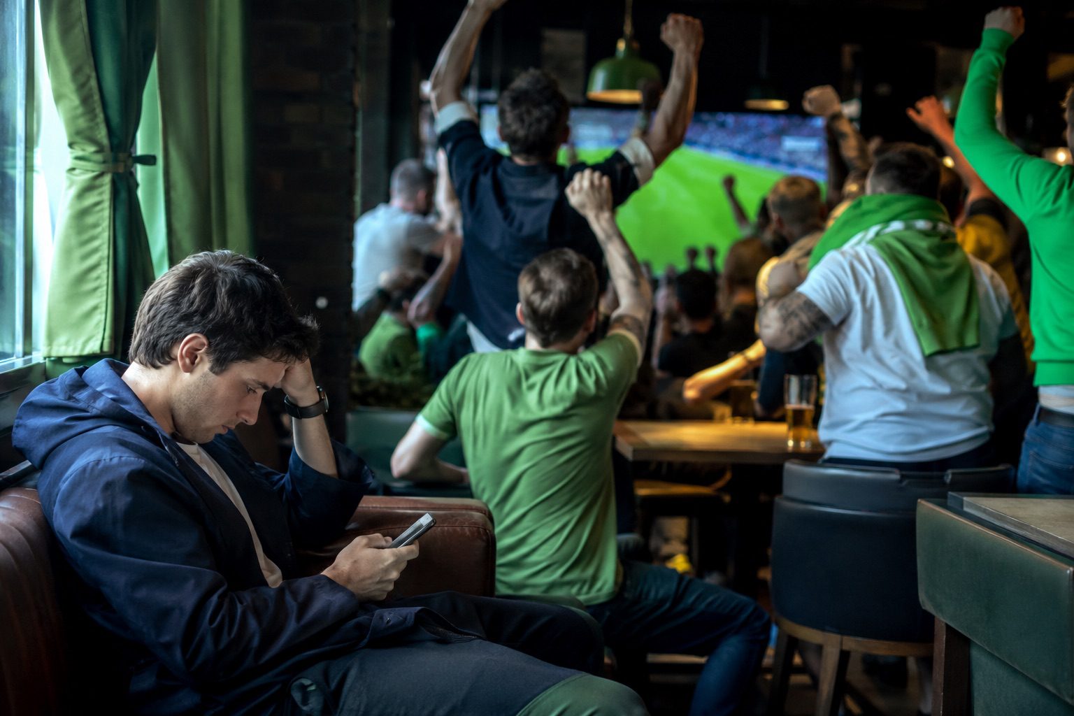 Man alone on his phone while fans behind him cheer in a sports bar